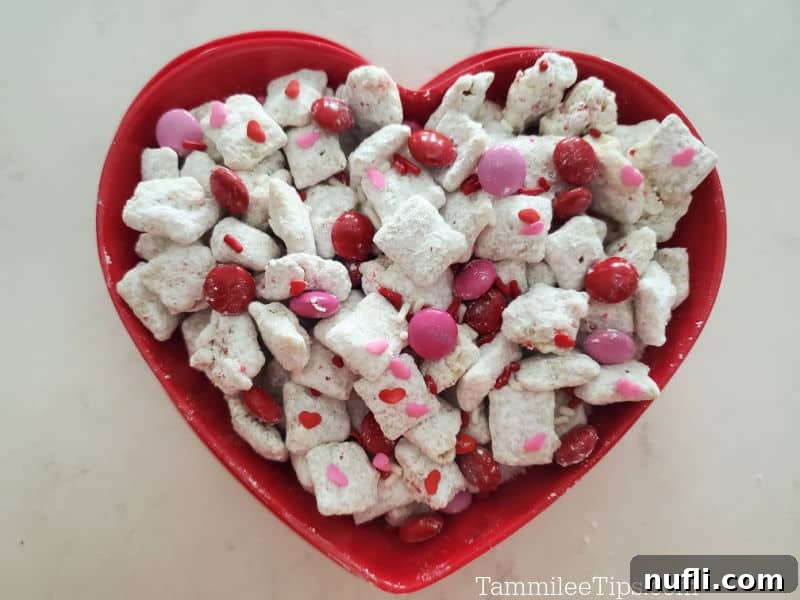 A red heart-shaped bowl brimming with Valentine's Day Muddy Buddies, featuring vibrant red and pink M&Ms.