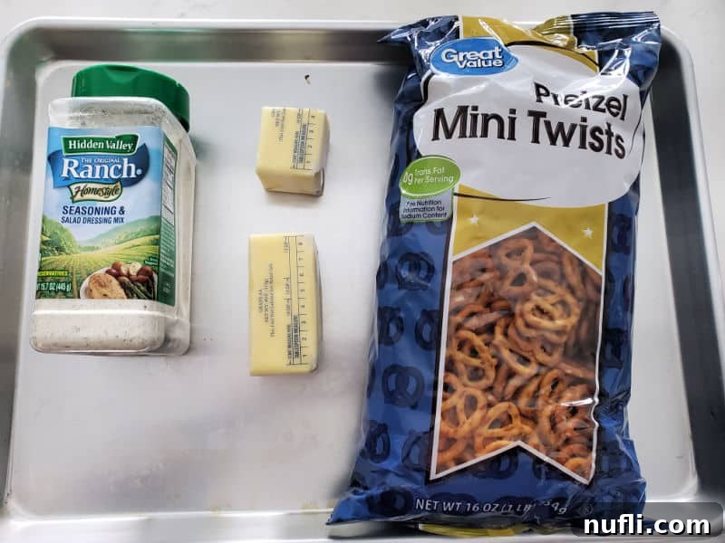 Essential ingredients for Ranch Pretzels: a container of dry ranch seasoning, a stick of butter, and a bag of mini pretzels on a baking sheet.