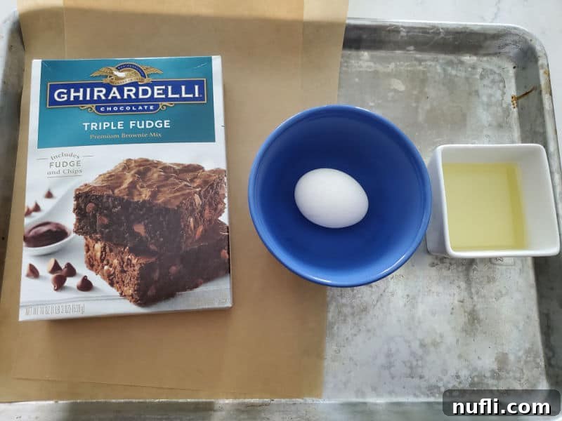 Ghirardelli triple fudge brownie box, egg in a blue bowl, and a bowl of oil on a baking sheet