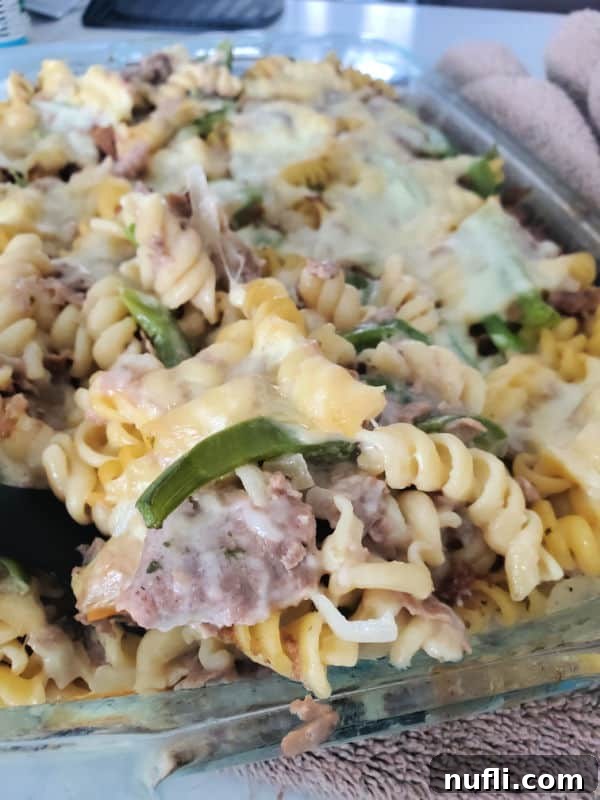 A serving spoon lifting a generous portion of cheesy Philly Cheesesteak Casserole out of a glass baking dish, showcasing the layers of pasta, steak, and melted cheese.