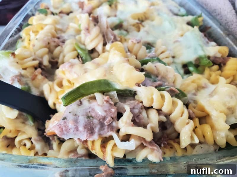 A spoon scooping creamy Philly Cheesesteak Casserole from a glass baking dish, highlighting the pasta and beef mixture.