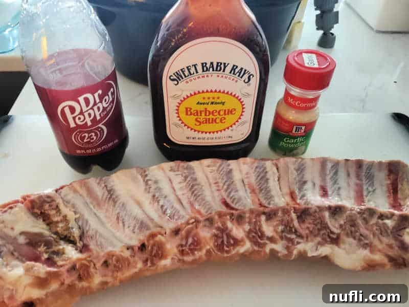 Ingredients for Dr. Pepper Crock Pot Ribs laid out on a cutting board: uncooked ribs, a bottle of Dr. Pepper, a bottle of barbecue sauce, and a small container of garlic powder.