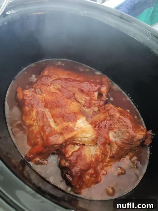 Close-up view inside a dark slow cooker, showing ribs fully covered in a rich, dark sauce, simmering gently.