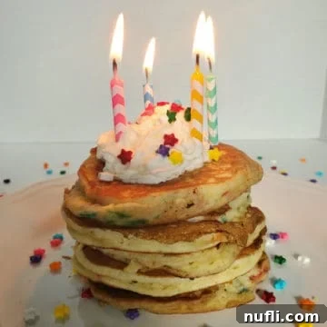 A beautifully arranged stack of Funfetti Cake Mix Pancakes on a white plate, complete with lit birthday candles, a generous dollop of whipped cream, and colorful sprinkles for a festive touch.