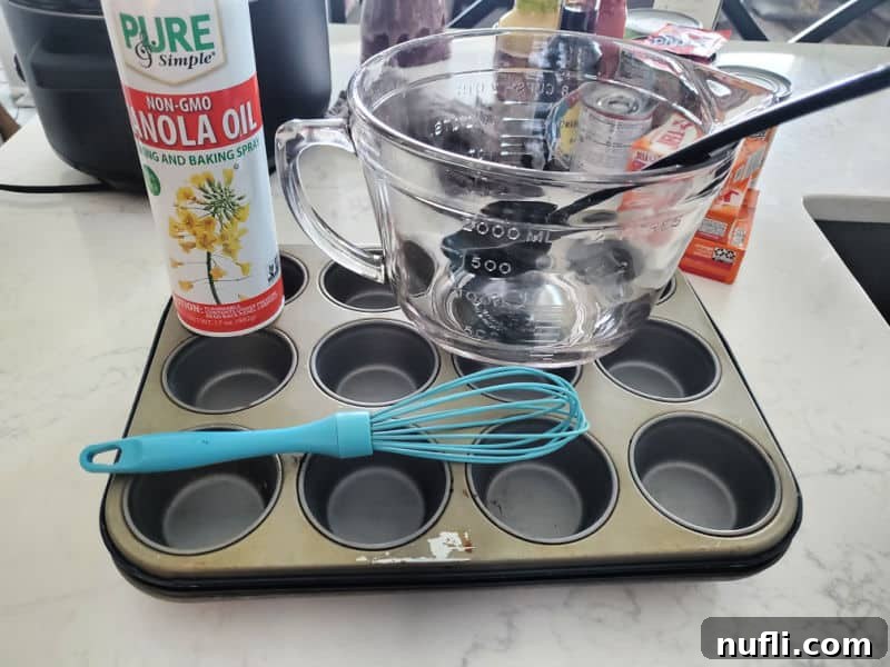 Various kitchen tools neatly arranged on a crowded marble counter, including a large mixing bowl, a whisk, a muffin tin, a can of canola oil spray, and a serving spoon, all ready for baking.