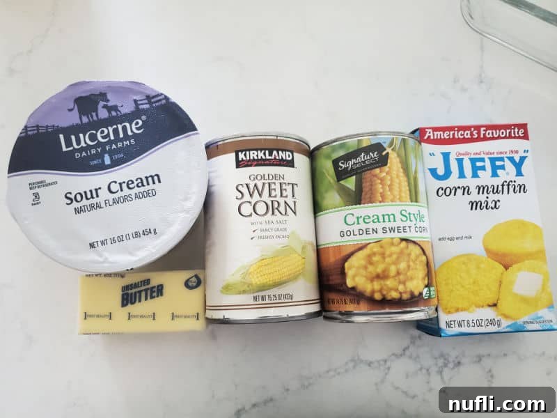 Ingredients for Creamed Corn Casserole laid out on a white countertop: sour cream, butter, sweet corn, golden style corn, and Jiffy corn muffin mix.
