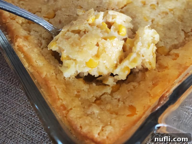A large serving spoon scooping out a generous portion of creamy, golden creamed corn casserole from a glass baking dish, ready to be enjoyed.