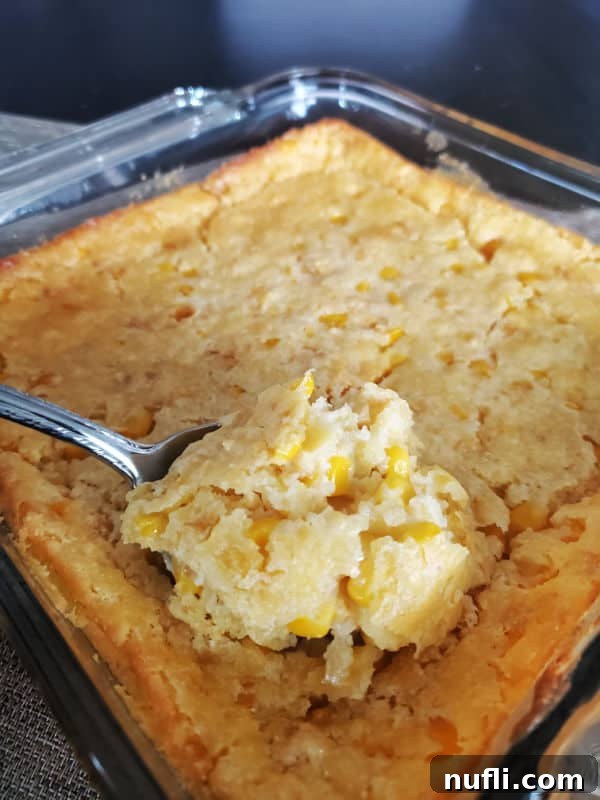 A large serving spoon holds a portion of creamy, baked creamed corn casserole, highlighting its delicious texture and golden-brown top, ready to be served from the baking dish.
