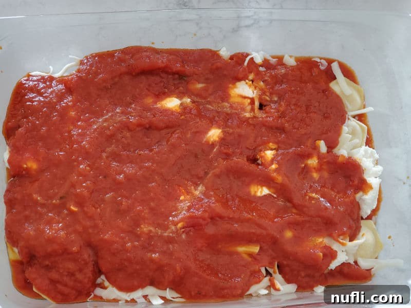Another layer of marinara sauce and shredded mozzarella cheese covering the ravioli in a glass casserole dish, building the lasagna layers.