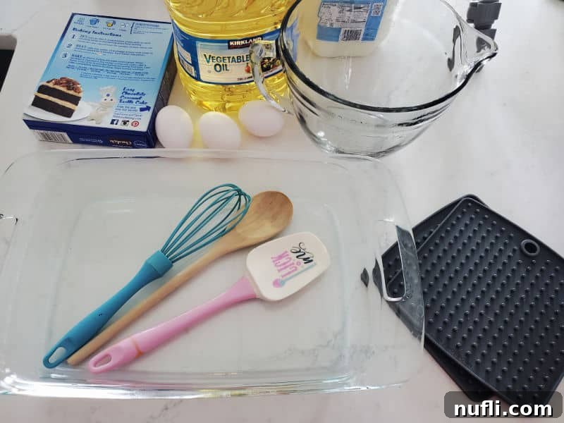Baking essentials for the Oreo Poke Cake, including a cake mix box, eggs, vegetable oil, a glass batter bowl with a whisk, a wooden spoon, a spatula, a glass baking dish, and pot holders on a white counter.