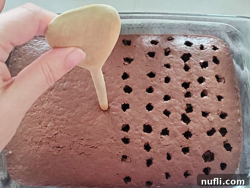 A wooden spoon being used to poke holes into a baked chocolate cake in a glass baking dish, preparing it for the pudding mixture.