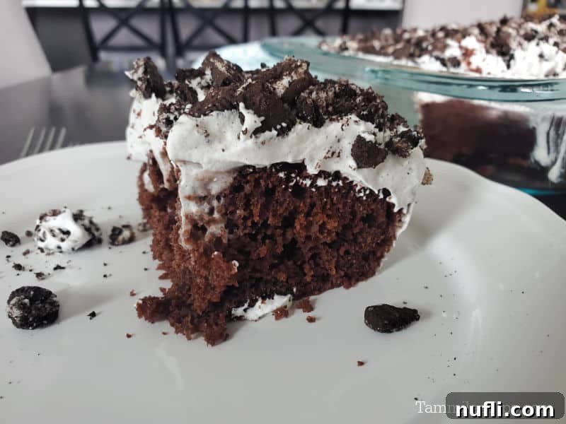 A square piece of Oreo Poke Cake on a white plate with the baking dish blurred in the background, showing the layers and crushed Oreo topping.