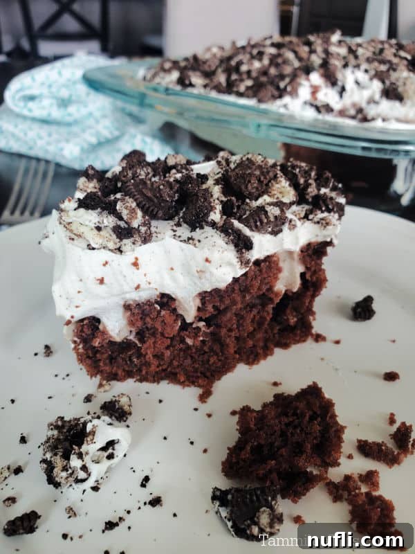 A slice of Oreo Poke Cake on a white plate, positioned next to the baking dish containing the rest of the cake.
