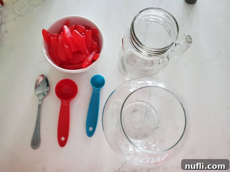 Homemade Sonic Ocean Water Drink Guide 6 A selection of items for serving Sonic Ocean Water, including Swedish Fish candy in a bowl, a mason jar glass, a decorative fishbowl, and measuring tools on a white counter.