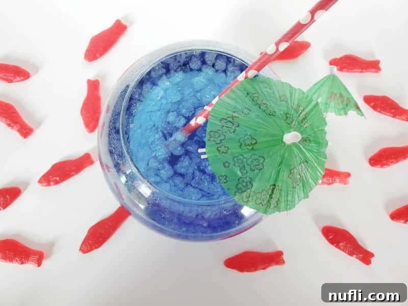 Homemade Sonic Ocean Water Drink Guide 9 A fish bowl filled with blue Sonic Ocean Water, garnished with a green umbrella and red straw, surrounded by colorful Swedish Fish Candies.