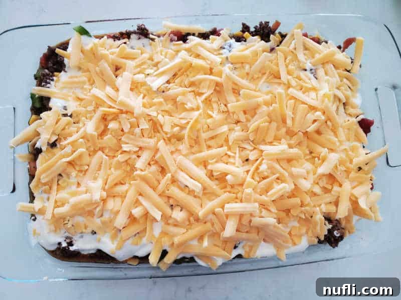shredded cheddar cheese over ground beef, sour cream, and vegetables in a casserole dish