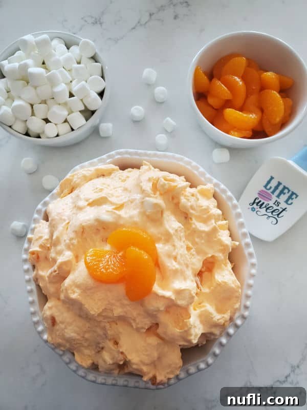 Orange jello salad with mandarin orange slices on top in a white bowl next to a bowl of mini marshmallows, a bowl of mandarin orange slices and a rubber spatula