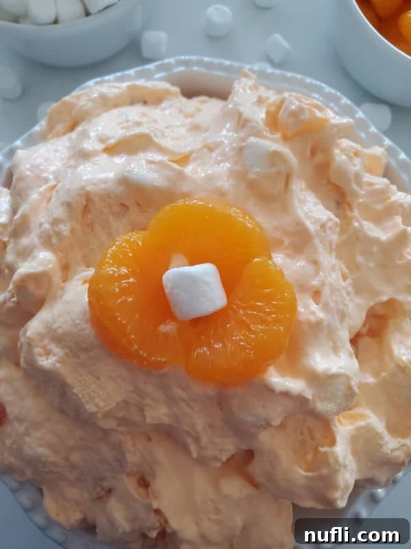 Close up of orange jello salad with three mandarin orange slices and a mini marshmallow on top in a white bowl 