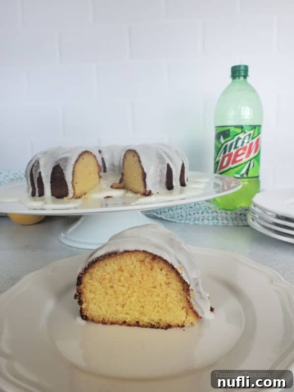A perfectly sliced piece of Mountain Dew cake on a white plate, positioned next to the remaining bundt cake on a platter and a bottle of Mountain Dew, highlighting its delicious texture and inviting presentation.