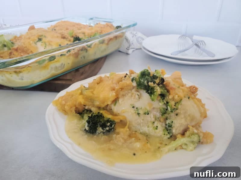 A close-up of a serving of broccoli cheddar chicken casserole on a white plate, with the baking dish blurred in the background, highlighting the creamy texture and crispy topping.