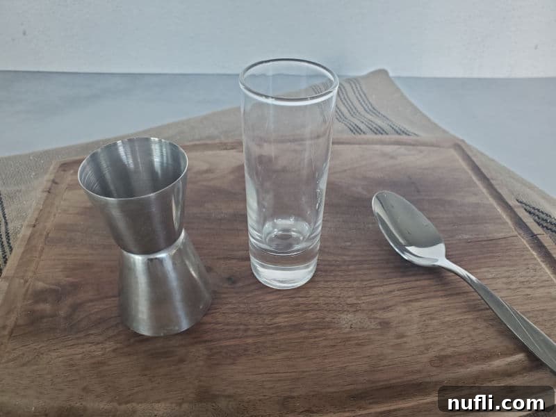 A jigger, shot glass, and bar spoon are neatly arranged on a wooden cutting board, essential tools for crafting the perfect layered shot.
