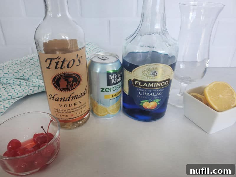 Ingredients for a Blue Lagoon Cocktail: a small bowl of maraschino cherries, a bottle of Tito's Vodka, a jug of lemonade, a bottle of Blue Curaçao, a hurricane glass, and lemon wheels, all arranged on a white counter.