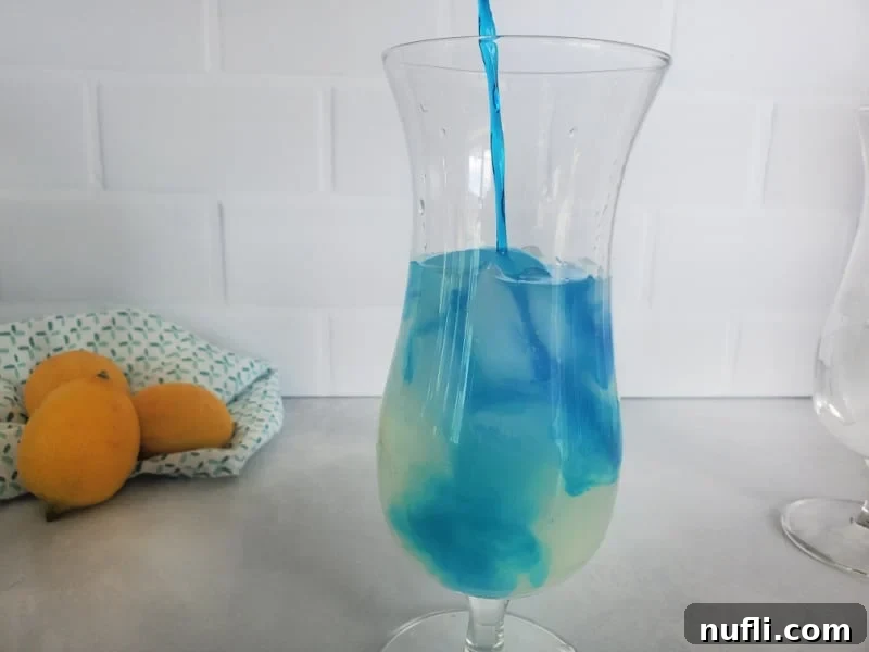 Blue Curaçao being poured into a hurricane glass over lemonade and ice, with lemons in the background.
