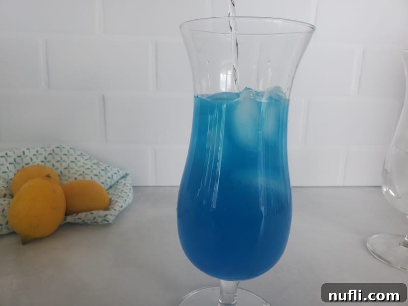 Clear vodka being poured into the blue liquid of a Blue Lagoon Cocktail in a hurricane glass, with lemons in the background.