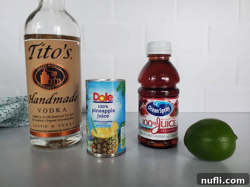 Bay Breeze Drink ingredients laid out: Titos Vodka bottle, a carton of Pineapple Juice, a bottle of Cranberry Juice, and a fresh lime.
