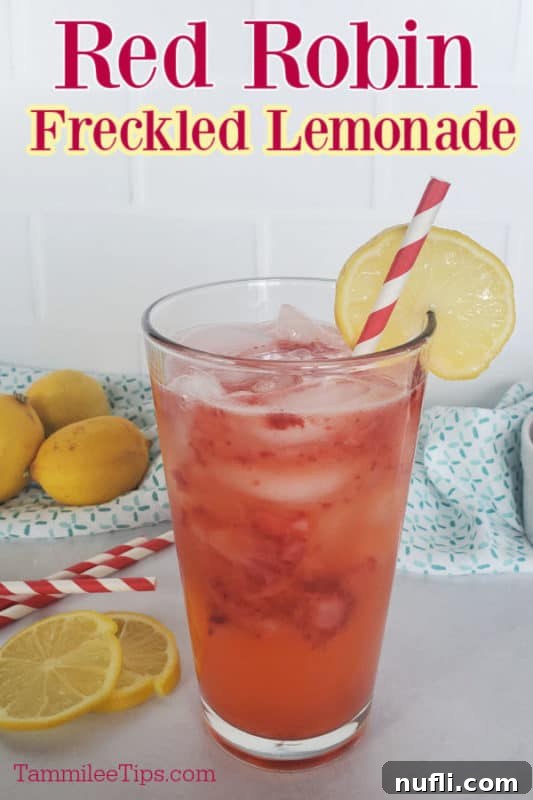 A tall glass of vibrant Red Robin Freckled Lemonade, garnished with a fresh lemon wheel and a strawberry. Whole lemons and a white cloth are visible in the soft-focus background, suggesting freshness and homemade goodness.
