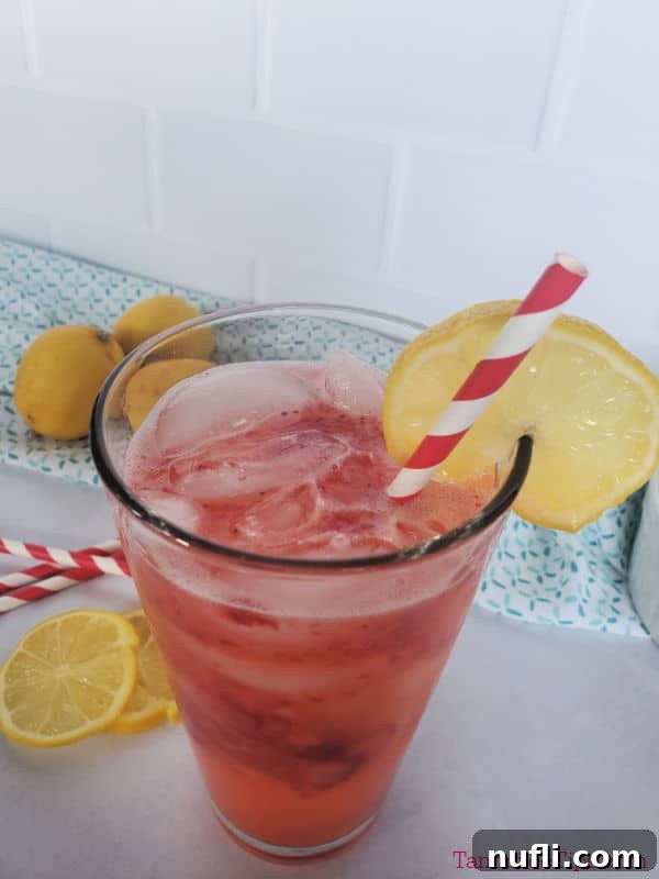 A refreshing glass of freckled lemonade with a bright lemon wheel garnish, placed on a white surface with blurred lemons and striped paper straws in the background, evoking a perfect summer day.