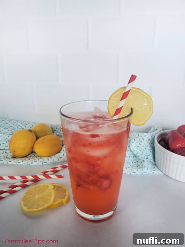 A close-up of a refreshing strawberry lemonade in a glass, featuring vibrant red strawberry pieces and a bright yellow lemon wheel garnish. The background is softly blurred with more lemons and striped straws, enhancing the inviting summer aesthetic.
