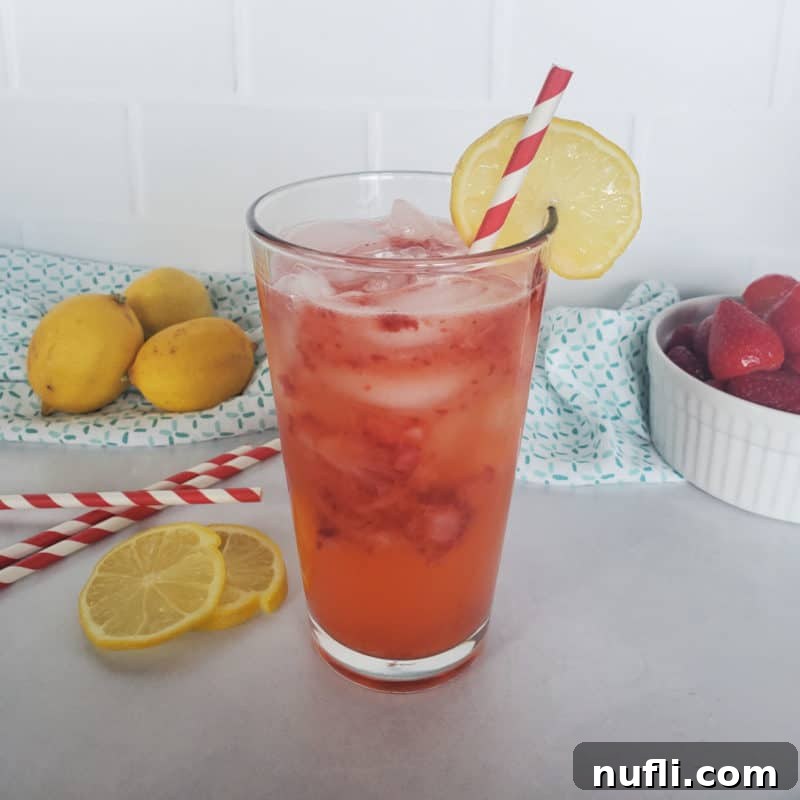 A vibrant glass of Freckled Lemonade, garnished with a lemon slice and showcasing the delightful crushed strawberries, set against a backdrop of fresh lemons and striped straws.