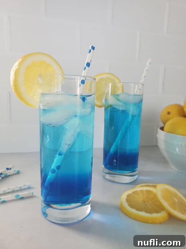 Two blue drinks in tall glasses garnished with lemon wheels and paper straws next to a bowl of lemons. 