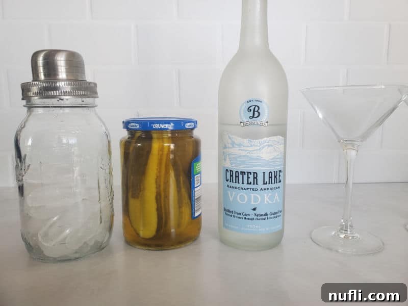 cocktail shaker, jar of pickles, bottle of vodka, and martini glass on a counter