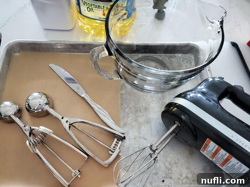Baking tools ready for making cookies: cookie scoops, a knife, parchment paper, a glass mixing bowl, a hand mixer, and baking sheets laid out on a kitchen counter.