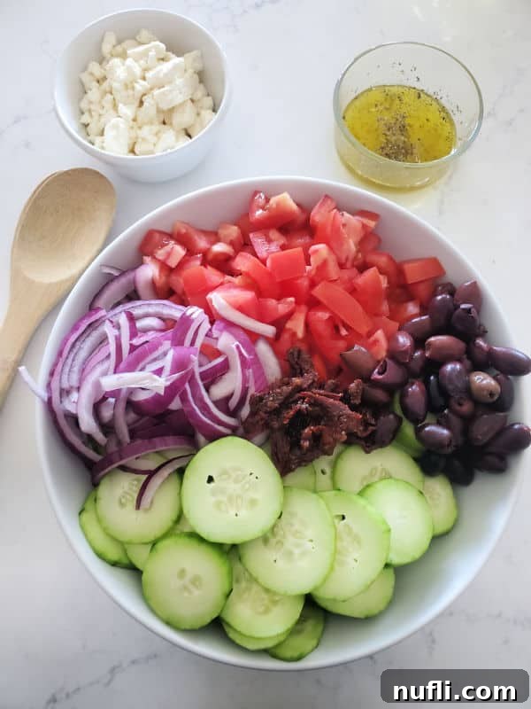 A large white bowl filled with chopped cucumbers, tomatoes, sun-dried tomatoes, and Kalamata olives, ready for dressing, next to separate bowls of olive oil dressing and crumbled feta cheese.