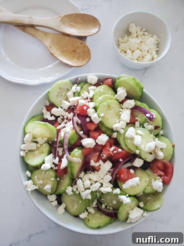 A refreshing Greek cucumber salad, topped with crumbled feta cheese, in a white bowl. Nearby, a plate with wooden serving spoons and an additional bowl of feta cheese are visible.
