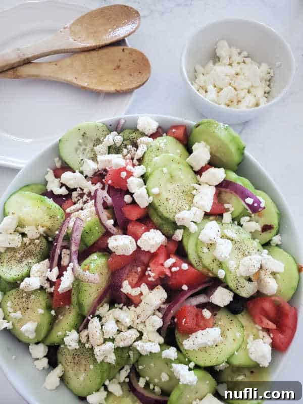 Close-up of Greek cucumber salad, lavishly topped with feta cheese, served in a white bowl. Wooden serving utensils and a bowl of extra feta are on the side.