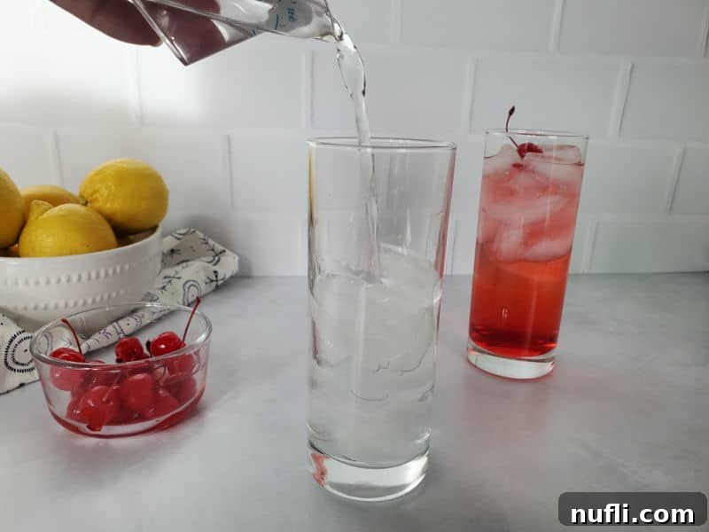 Clear vodka being poured into a tall glass filled with ice, next to a bowl of lemons and maraschino cherries.