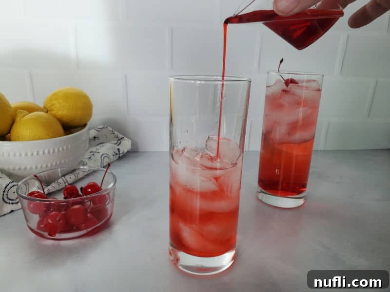 Red grenadine being poured into a tall glass containing ice and vodka, alongside a prepared Dirty Shirley cocktail.