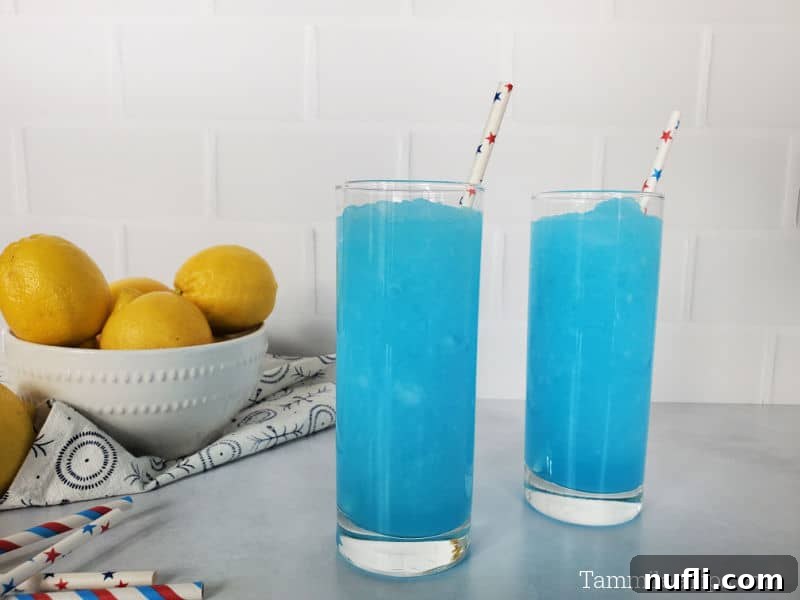 two bright blue kool aid slushies in tall glasses with paper straws, near a bowl of lemons and a stack of straws
