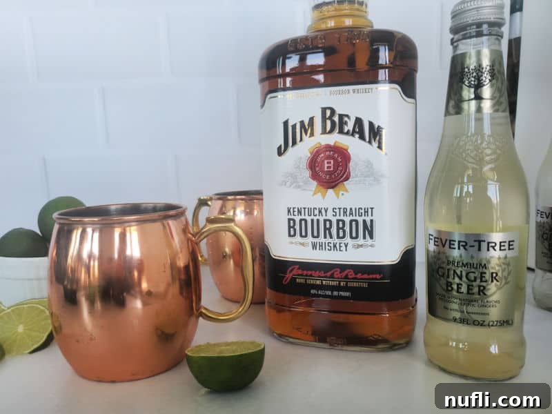The Best Easy Kentucky Mule Recipe 3 Key ingredients for a Kentucky Mule arranged on a surface: a shiny copper mug, a bottle of Jim Beam bourbon, several fresh limes, and a bottle of Fever-Tree ginger beer.