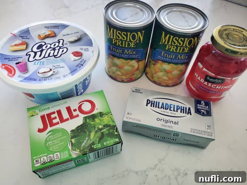 green jello salad ingredients on a marble counter with cool whip, two cans of fruit mix, philadelphia cream cheese and a packet of lime jello