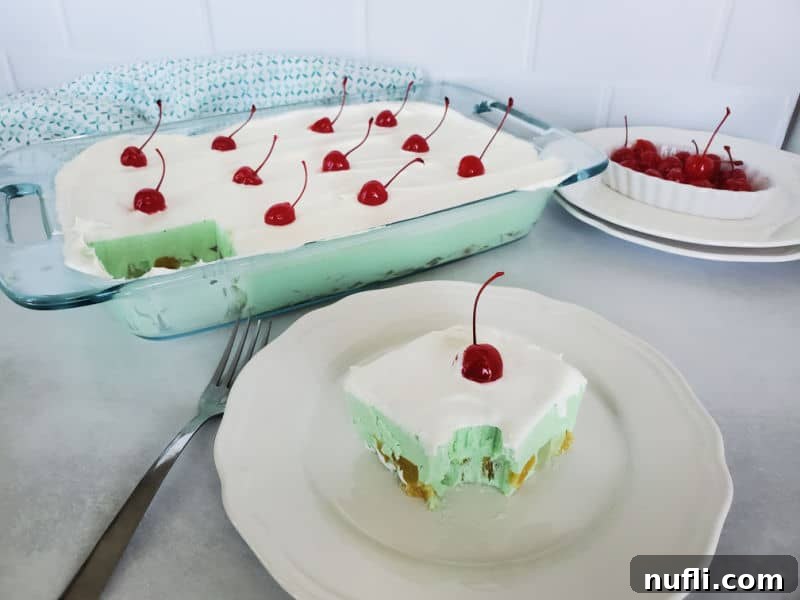 square of green jellow with cool whip and a maraschino cherry on a plate next to the baking dish and a bowl of cherries. 