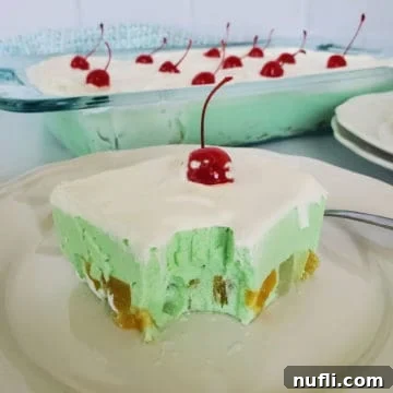 Square of green jello salad garnished with a cherry on a white plate next to a baking dish filled