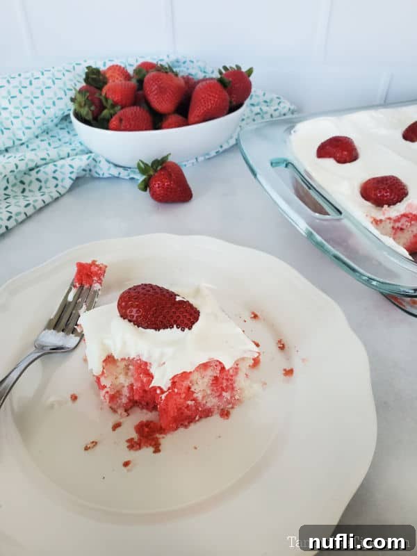 Slice of strawberry cake on a white plate with a fork next to a bowl of strawberries with a baking dish with cake in the corner