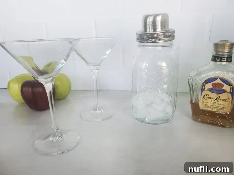 Red and green apples next to two glass martini glasses, a cocktail shaker with ice, and a bottle of Crown Royal
