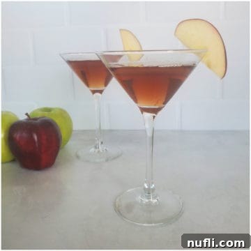 Two red brown martinis with apple slices in a martini glass next to green and red apples