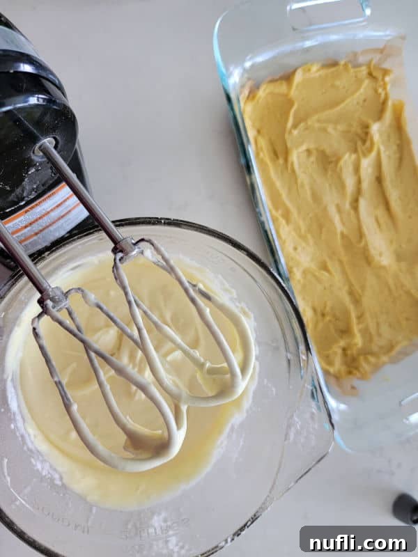 A handheld mixer hovering over a bowl of cream cheese topping mixture, with the cake base layer visible beside it.